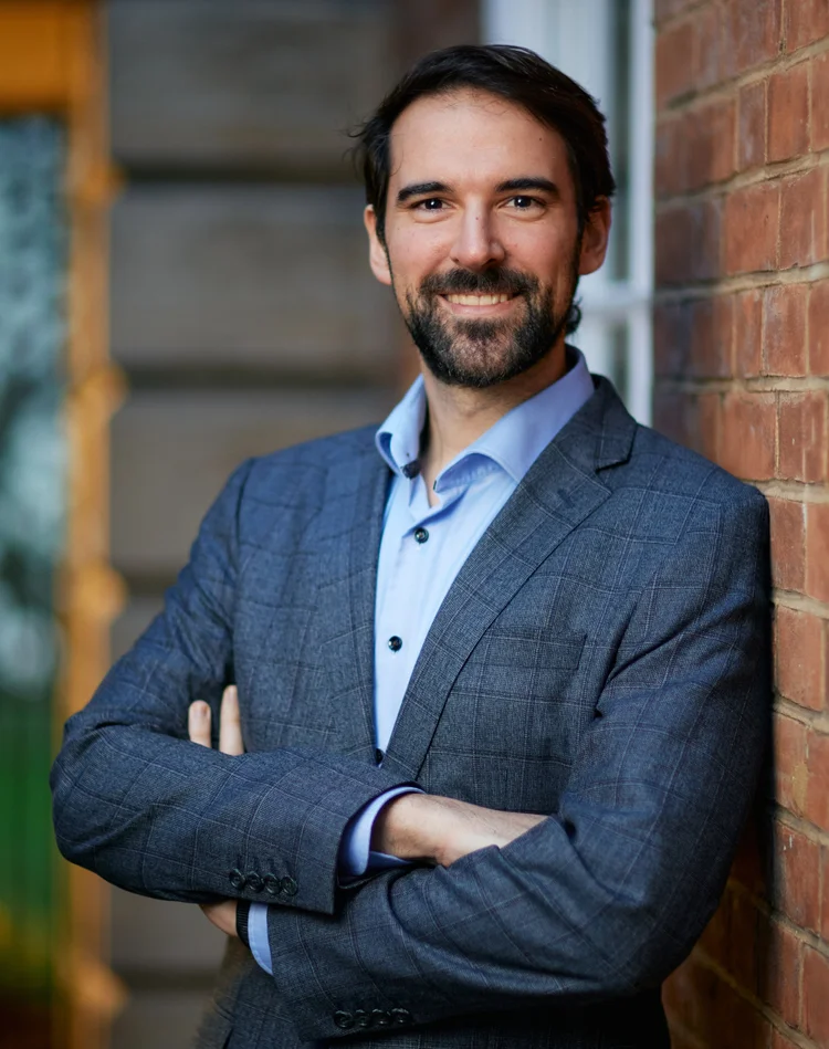A man with dark hair and a beard, wearing a blue dress shirt and a dark gray blazer, smiling with arms crossed against a brick wall.