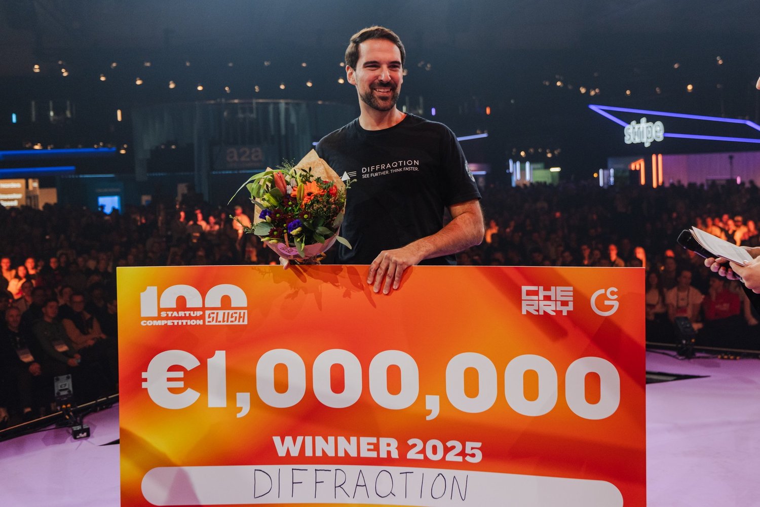 A man holding a bouquet of flowers and a large check for 1,000,000 euros at a startup competition event, with a crowd in the background and the word 'winner' and the year 2025 on the check.