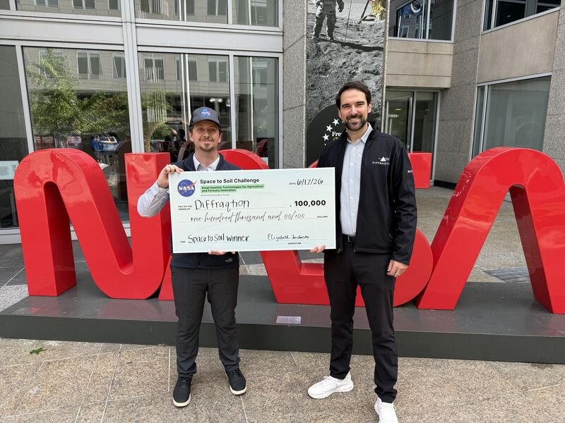 Two young men standing outdoors in front of a large red NASA sign, holding a large check awarded for the Space to Soil Challenge. One man wears a cap and the other a jacket, both smiling. The background includes glass windows and a mural on the wall.