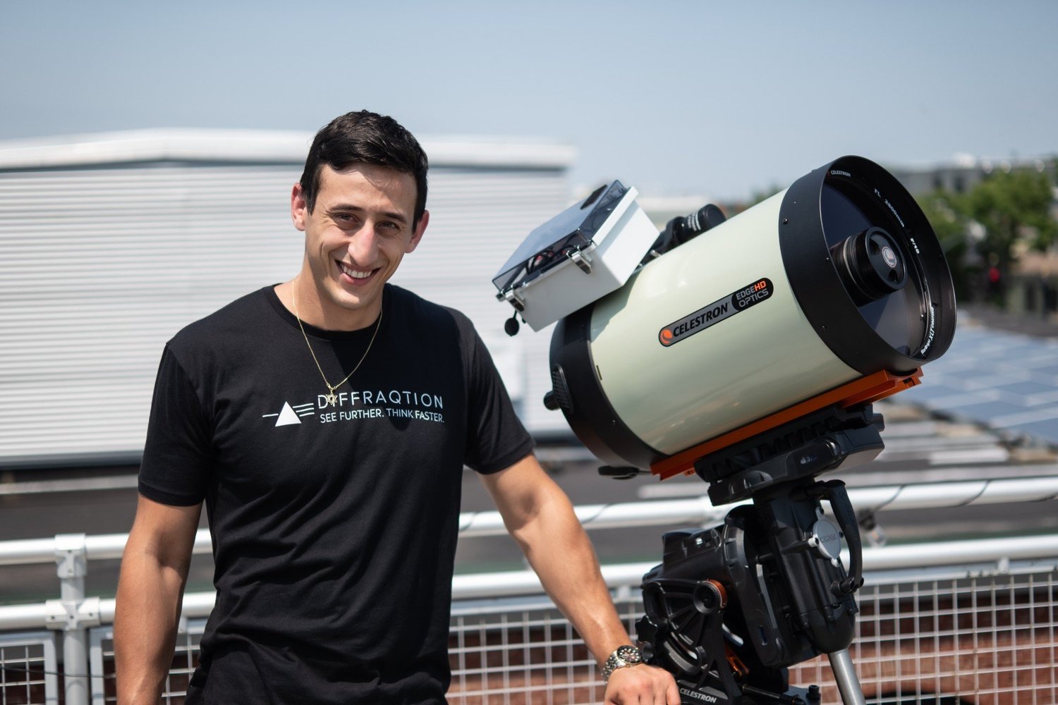 A smiling man standing next to a large telescope outdoors on a rooftop, with a white building and greenery in the background.