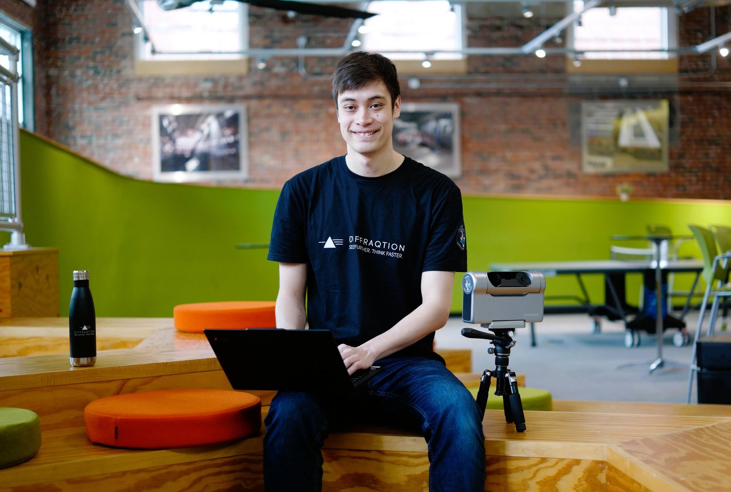 Young man sitting at a wooden table in a modern, industrial-style room, smiling at the camera. He is wearing a black t-shirt with a logo. On the table are a black water bottle, a laptop, and a tablet on a tripod. The background features brick walls, large windows, and colorful chairs.